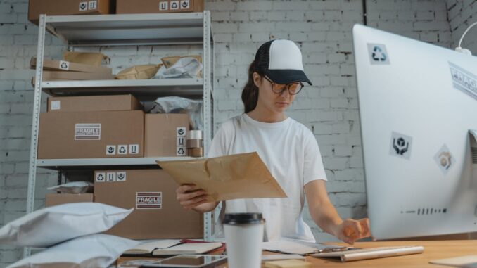 Kobieta w czapce i okularach pakuje paczki w małym magazynie e‑commerce; jest prawdopodone, że w takiej firmie wykorzystuje się najprostszy program magazynowy.