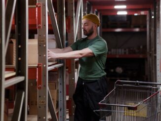 Mężczyzna w zielonej koszulce i czapce pracuje w magazynie, układając pudełka na regale obok wózka – ilustracja artykułu o codziennych obowiązkach logistyka.