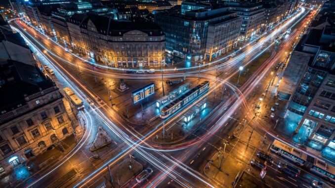 Nocne ujęcie z lotu ptaka na duże skrzyżowanie w centrum miasta, po którym pędzą samochody, tramwaje i autobusy, zostawiając świetlne smugi; wśród gęstej zabudowy widać przystanki i tablice informacji pasażerskiej, co symbolizuje miasto zarządzane algorytmami AI, które uczą transport wozić ludzi mądrzej.
