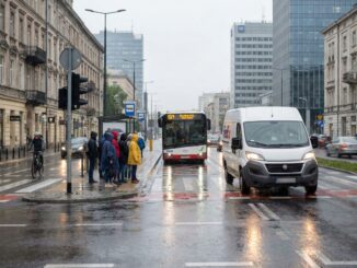 Deszczowa ulica w centrum miasta: autobus podjeżdża na przystanek, a obok jedzie furgonetka kurierska – codzienna logistyka dostaw w tle ruchu miejskiego.