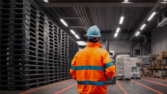 Pracownik w kamizelce odblaskowej w hali magazynowej – czerwone linie bezpieczeństwa z projektora liniowego LED wyznaczają ciągi komunikacyjne.