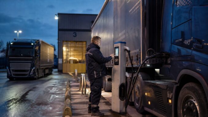 Elektryczny ciągnik siodłowy podłączony do ładowarki dużej mocy na placu bazy transportowej o zmierzchu.
